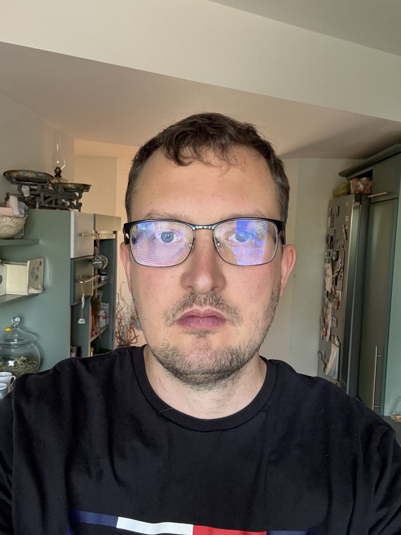 Selfie muže s brýlemi a černým tričkem v domácím interiéru.