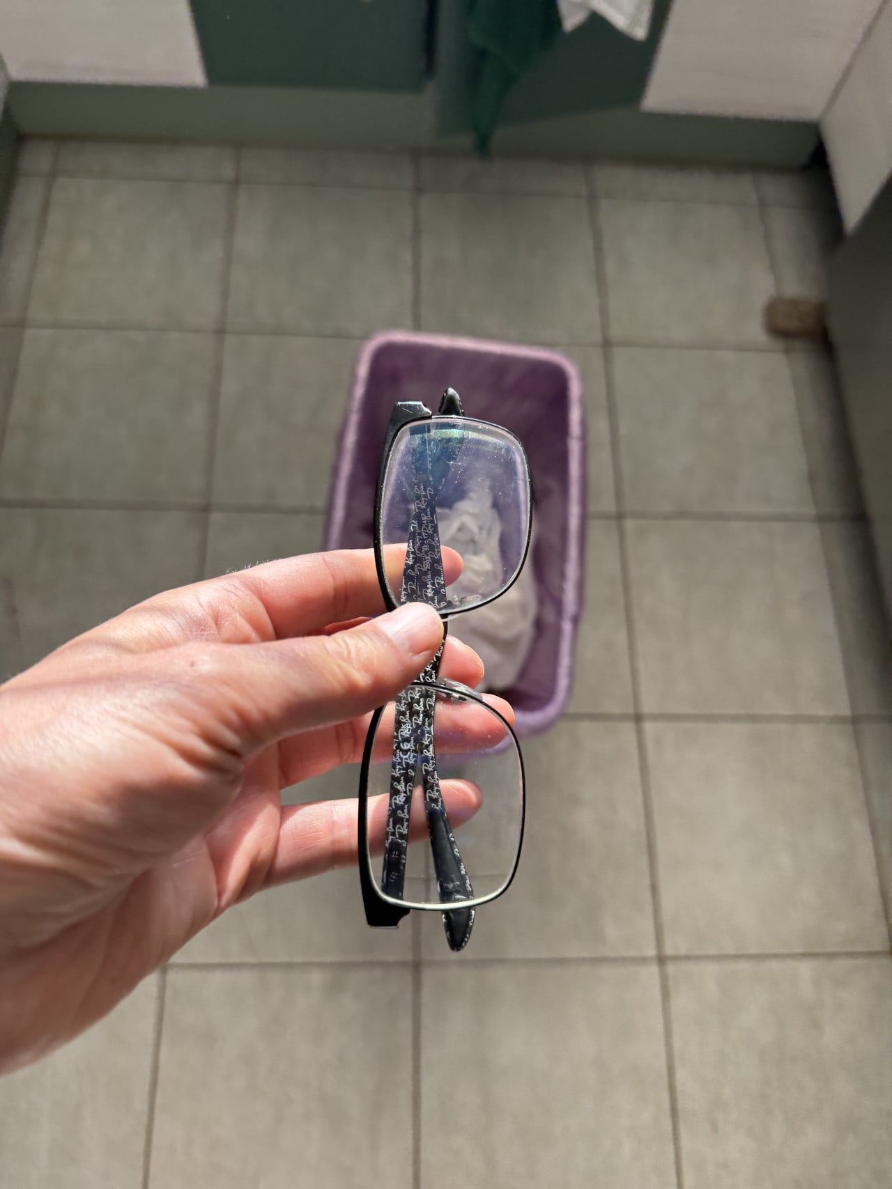 Ruka drží brýle nad košem v koupelně na dlaždicové podlaze.