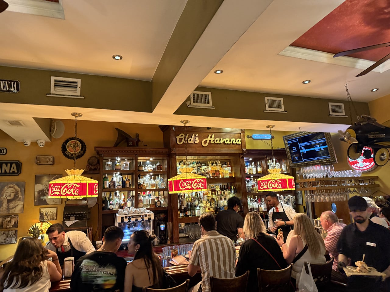Plný bar v Little Havaně, nápis Old's Havana, lampy Coca-Cola, hosté.