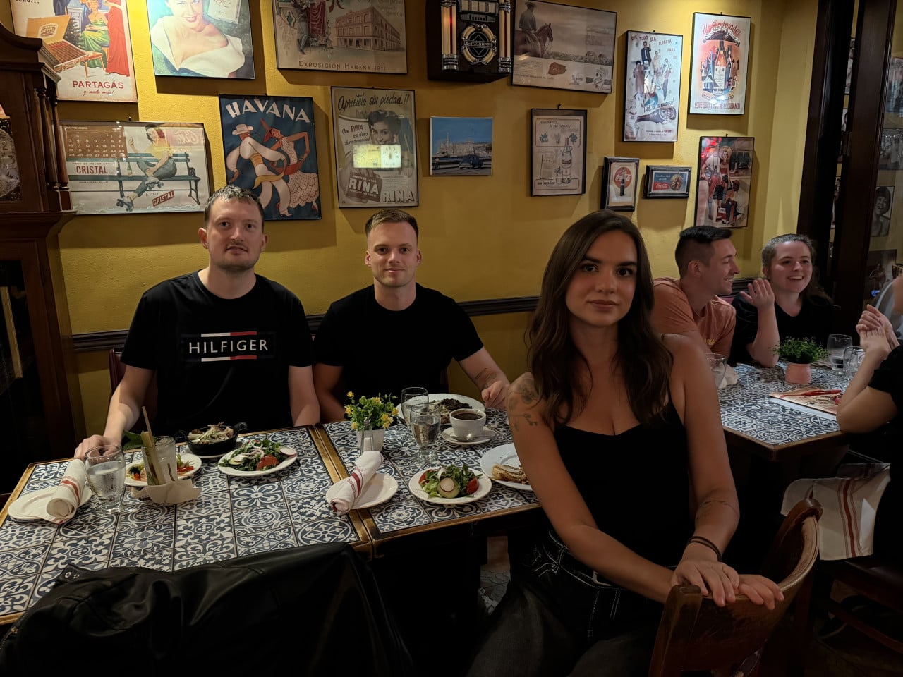 Tři lidé u stolu v kubánské restauraci, na zdi plakáty HAVANA, HILFIGER