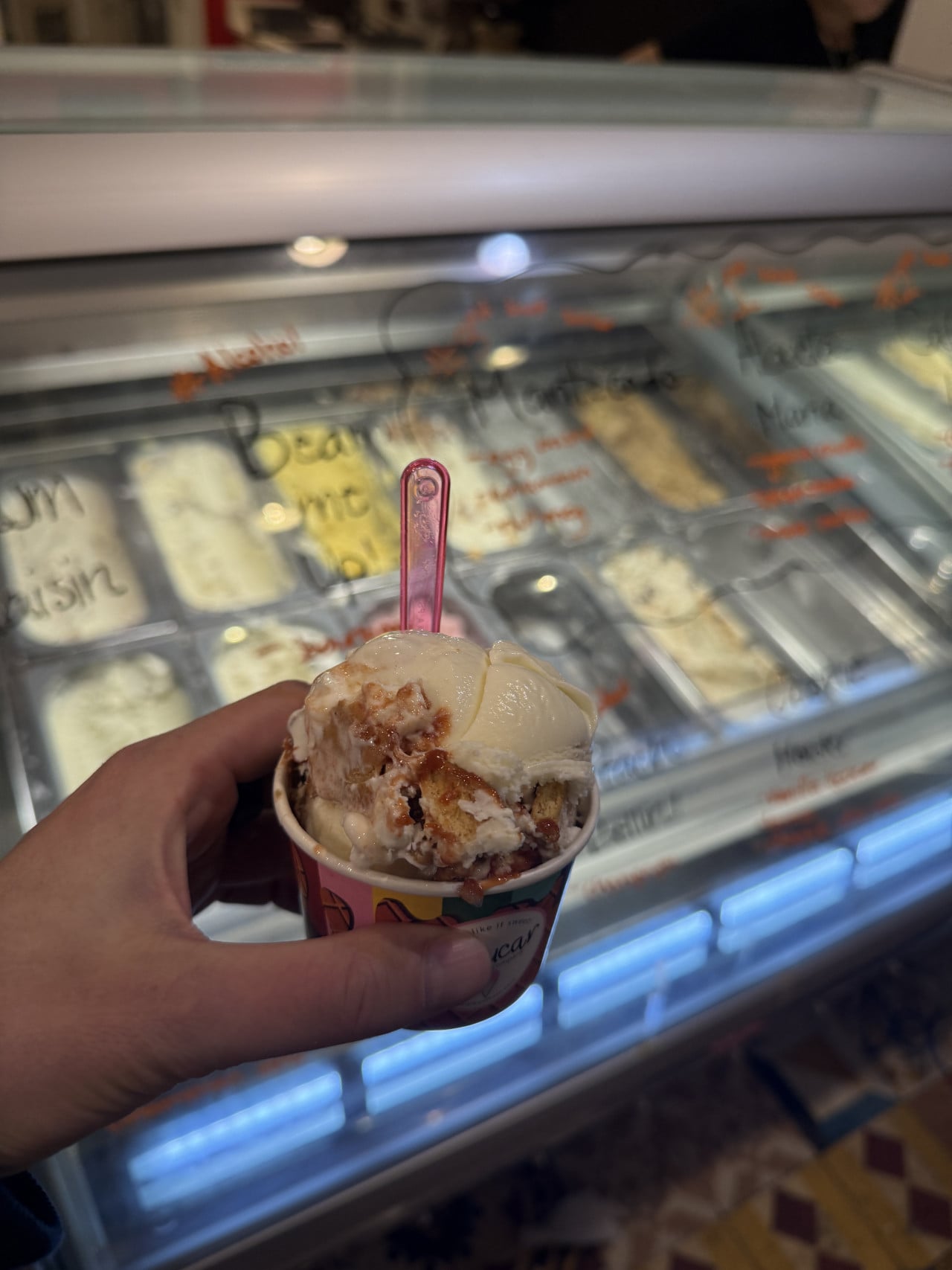 Ruka drží kelímek zmrzliny se lžičkou před vitrínou s příchutěmi