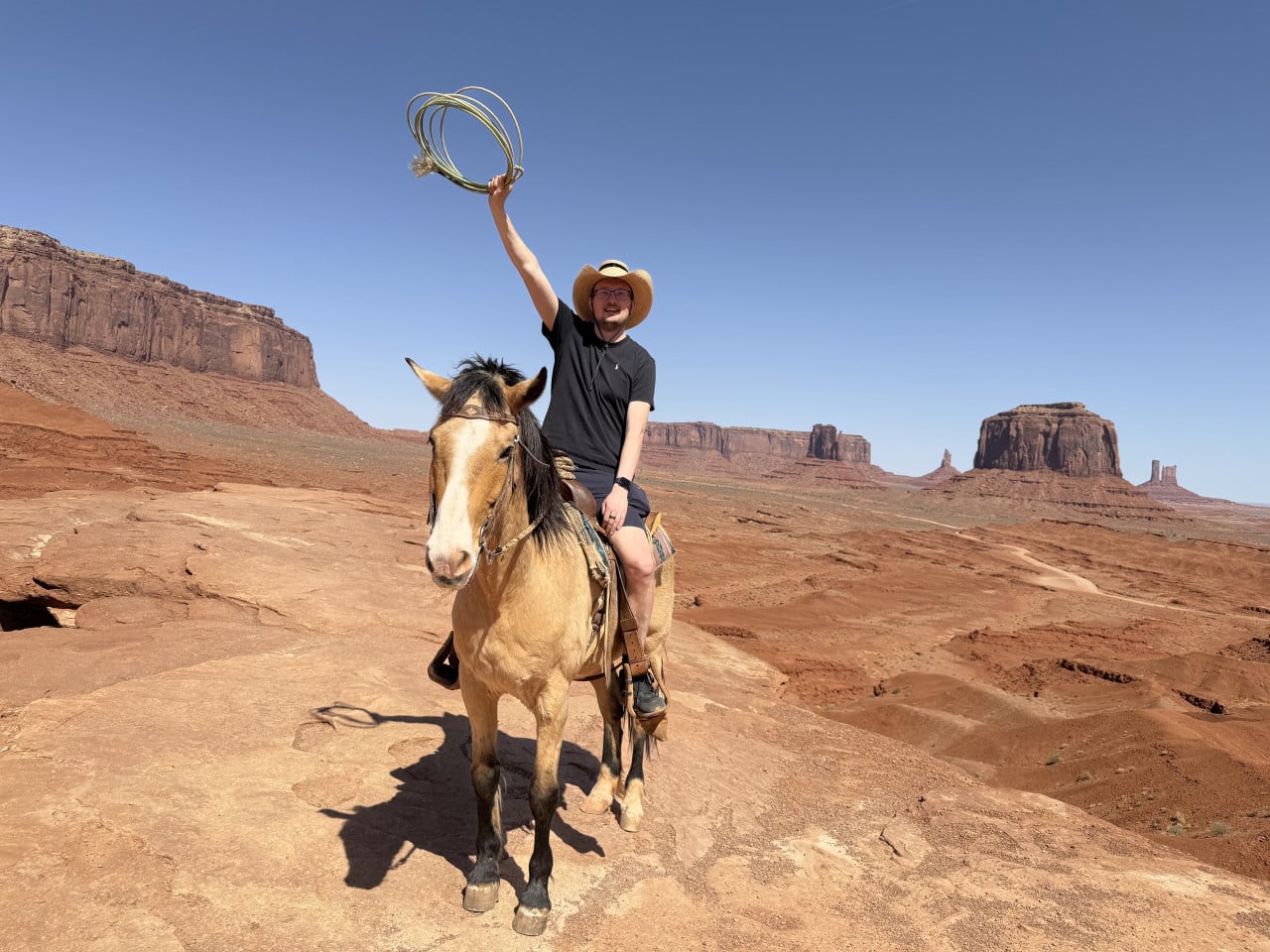 Muž na koni mává lasem, v pozadí skalní monumenty Monument Valley.