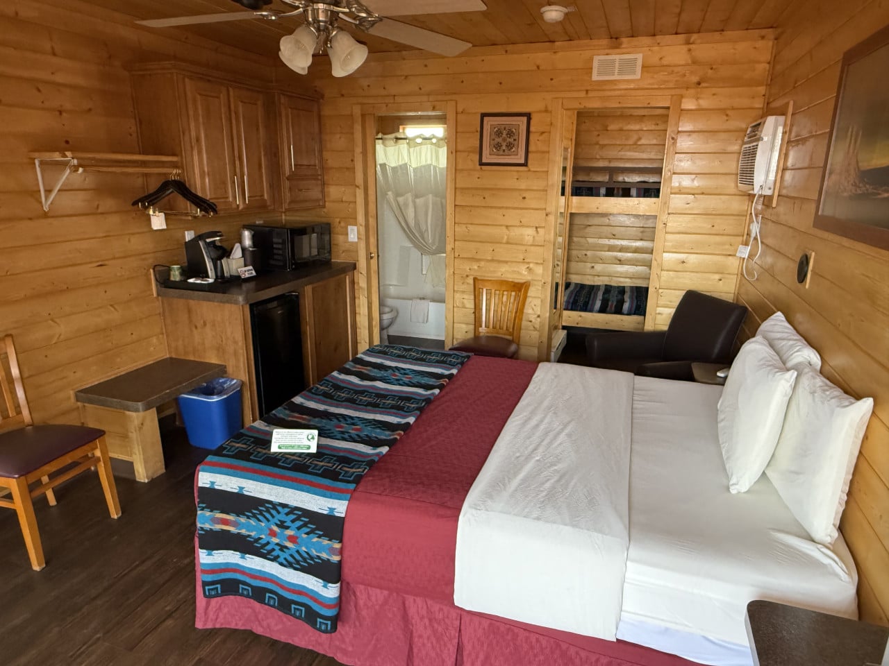 Interiér dřevěné chatky: dvojlůžko s dekou, kuchyňský kout, palanda a koupelna.