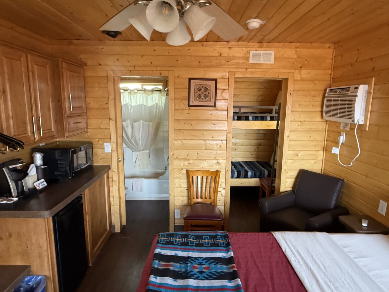 Interiér dřevěné chatky: postel, kuchyňka s mikrovlnkou, palanda, křeslo, koupelna.