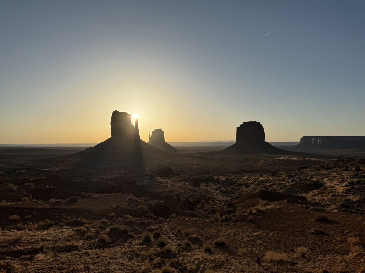 Východ slunce nad Monument Valley, slunce za skalními věžemi, poušť a kondenzační stopa