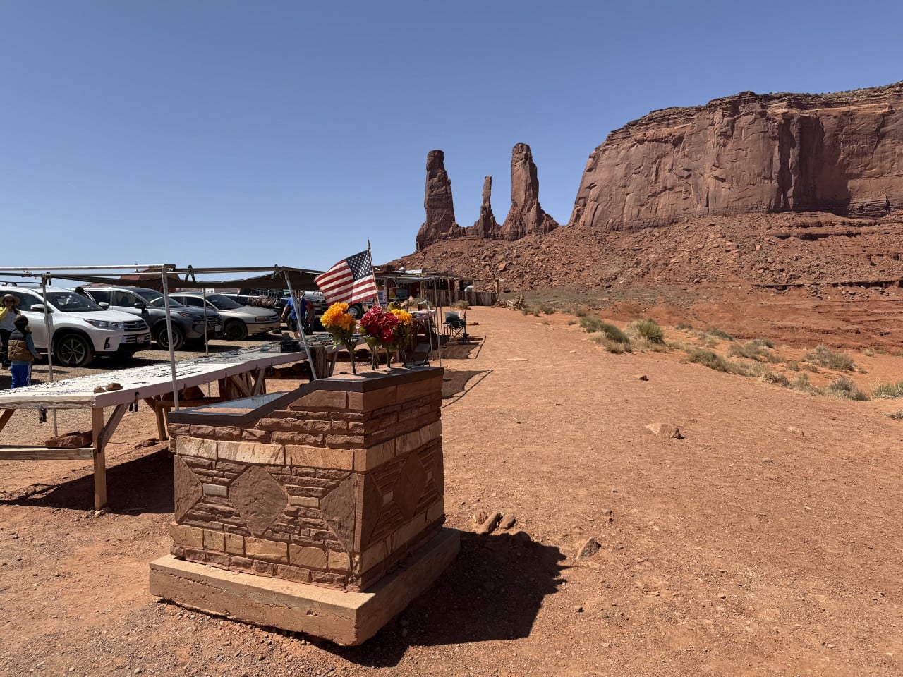 Americká vlajka a květiny u parkoviště, v pozadí skalní věže Monument Valley