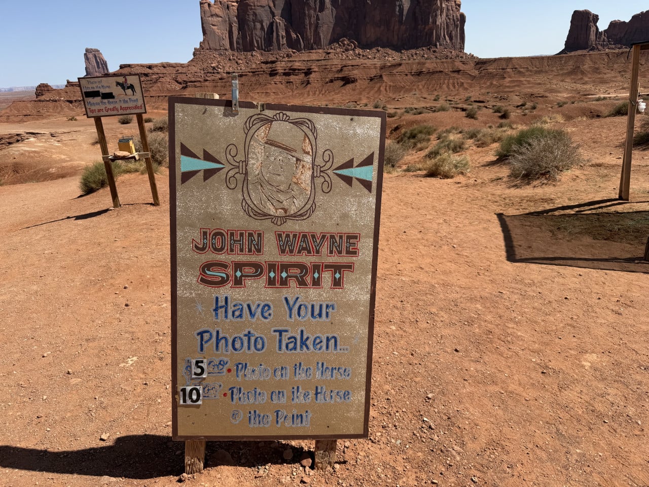 Cedule JOHN WAYNE SPIRIT Have Your Photo Taken, v pozadí monumenty Monument Valley.