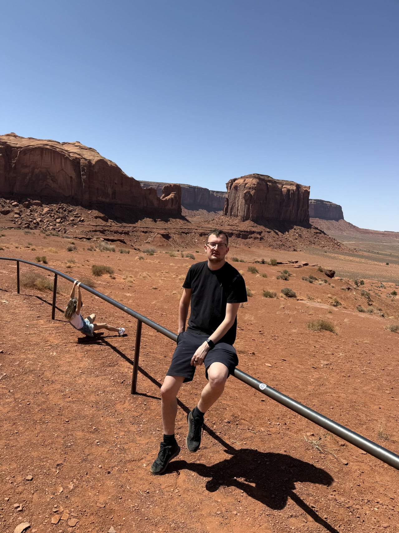 Muž sedí na zábradlí, dítě se houpe vedle, v pozadí skalní monumenty Monument Valley