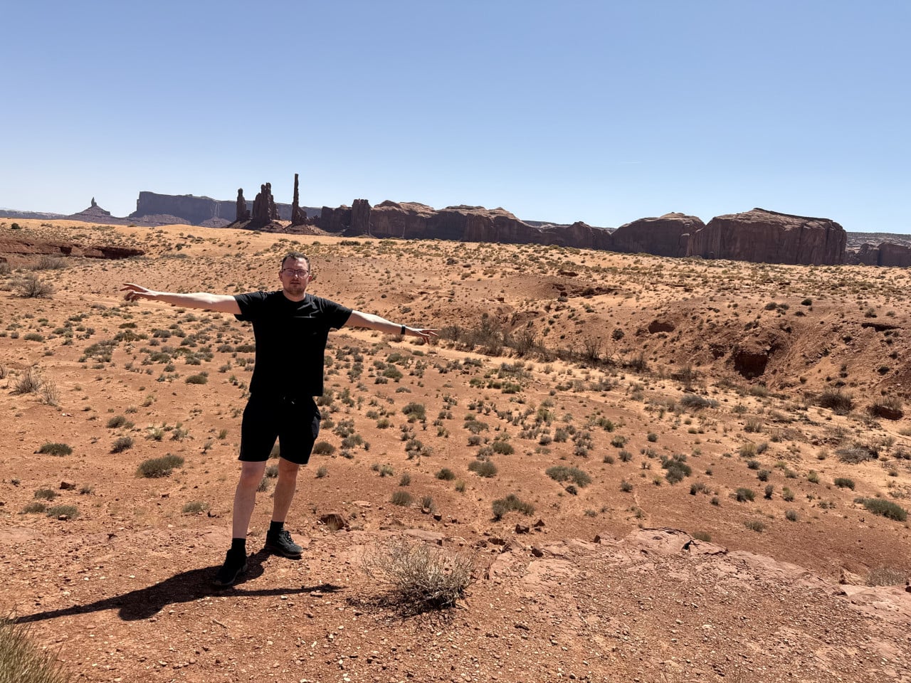 Muž v černém s rozpaženýma rukama na poušti, v pozadí skalní monumenty