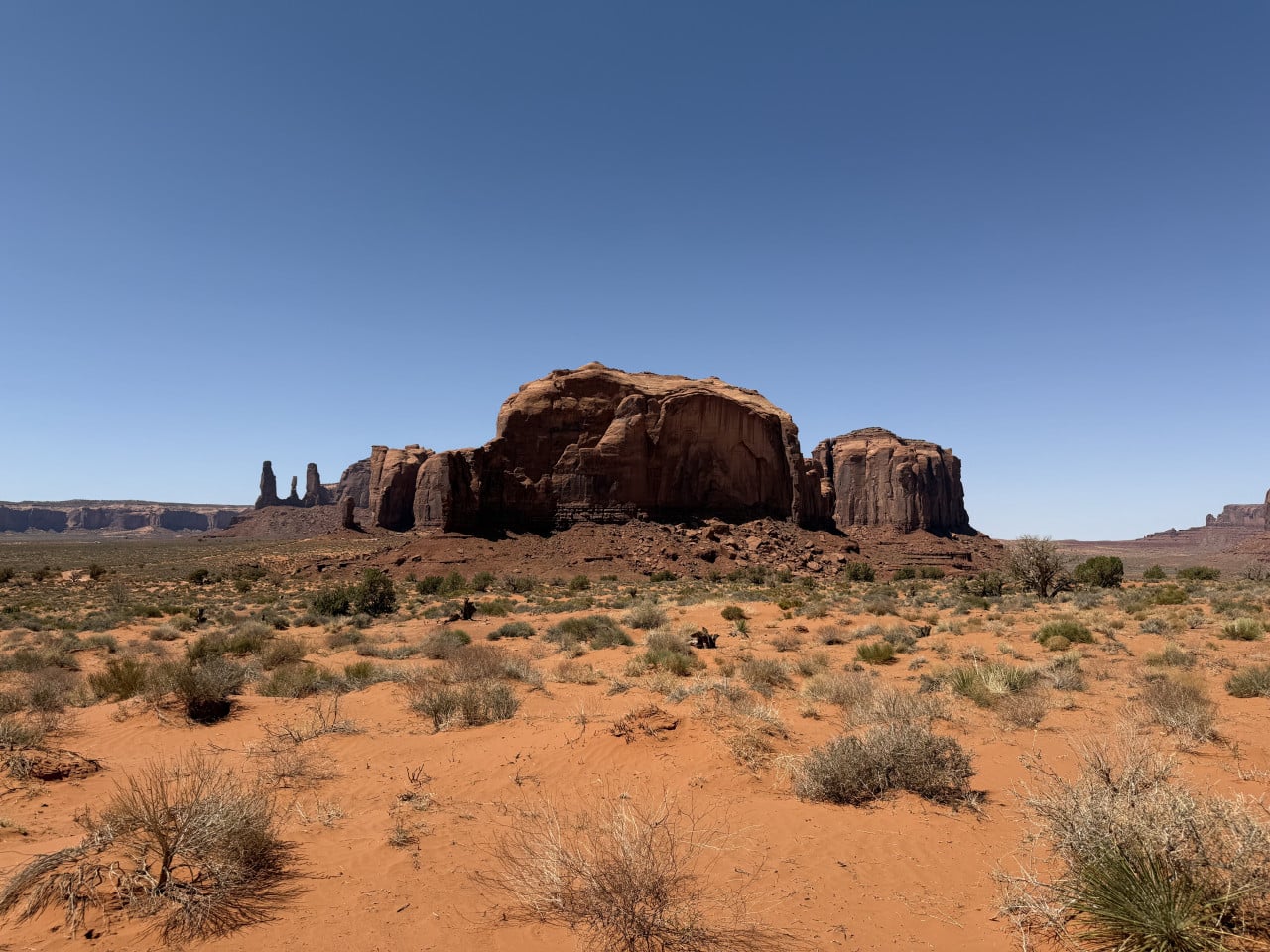 Pohled na skalní monumenty Monument Valley, červený písek, keře a modrá obloha