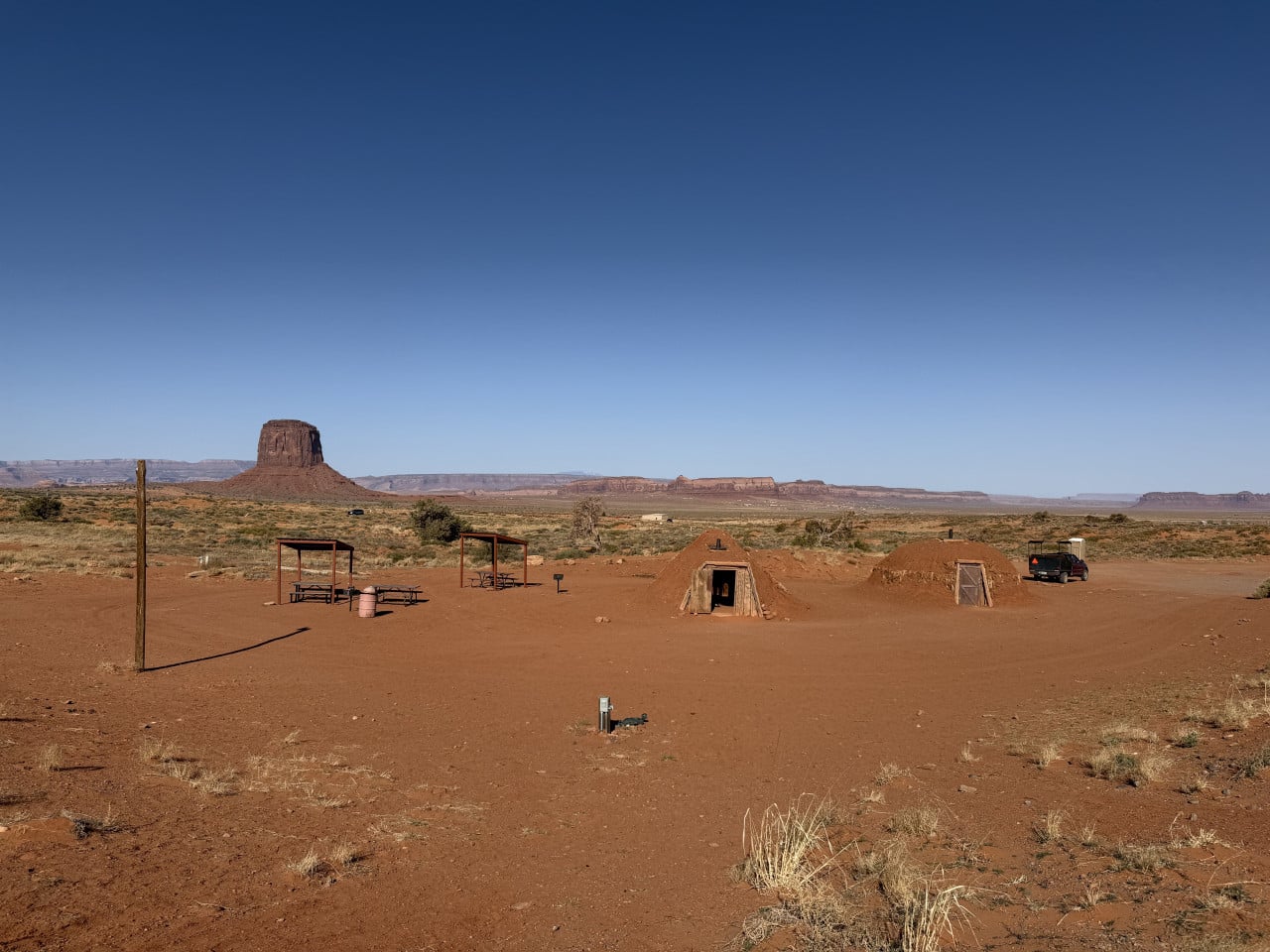 Kemp na červené půdě s chatkami, přístřešky a pickupem; v dálce monument