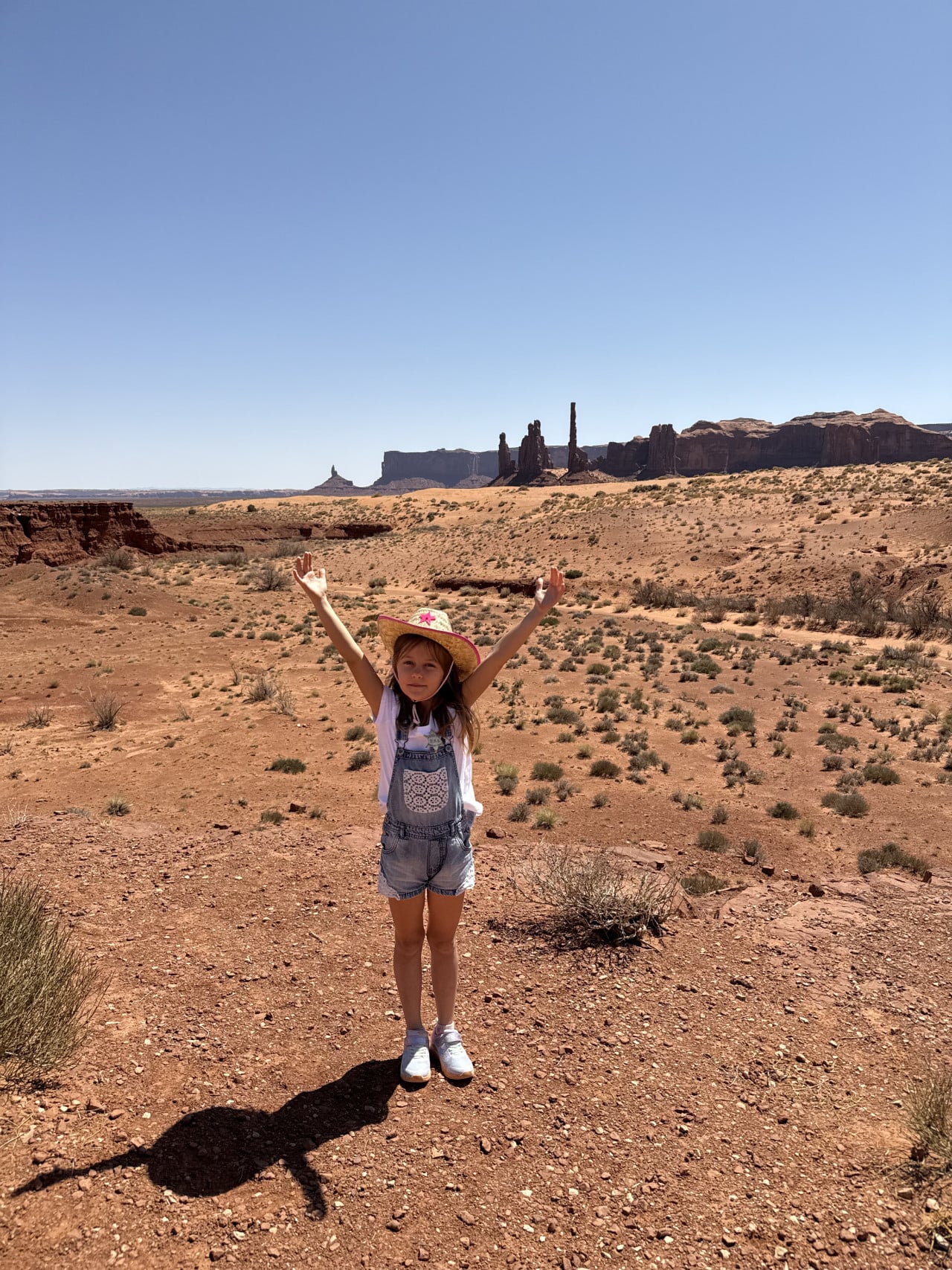 Dívka s kloboukem a zvednutýma rukama na poušti, v pozadí monumenty Monument Valley