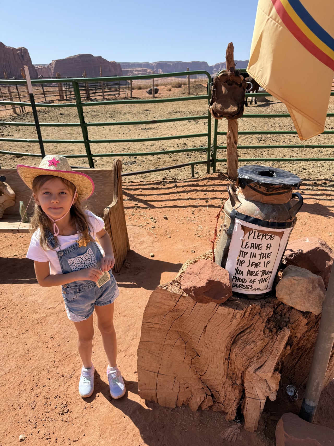 Dívka v klobouku u ohrady; cedule PLEASE LEAVE A TIP, v pozadí Monument Valley.