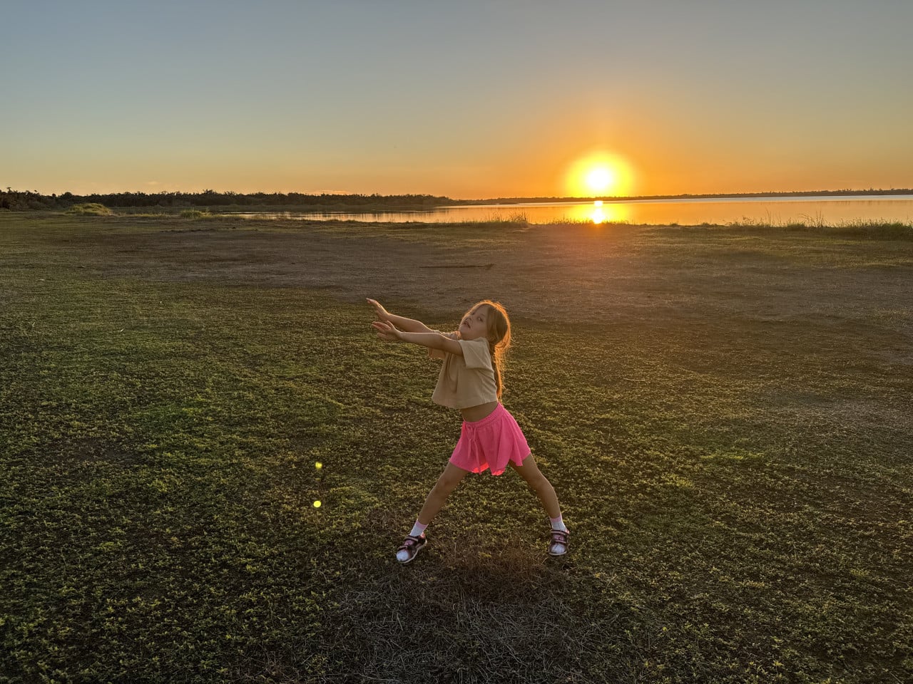 Dívka v růžové sukni pózuje na louce při západu slunce u jezera.