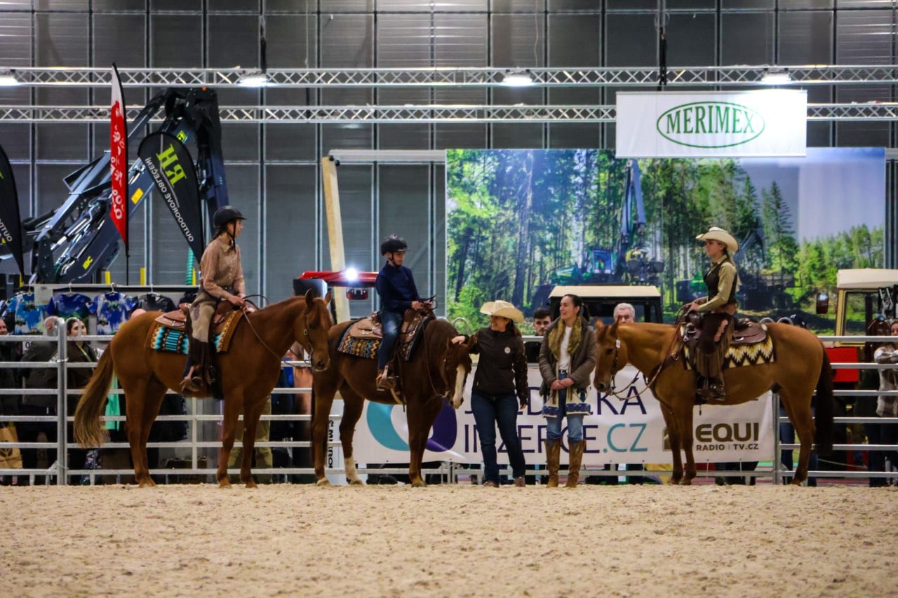 Tři jezdci na koních v aréně, kolem stojí diváci a bannery MERIMEX.