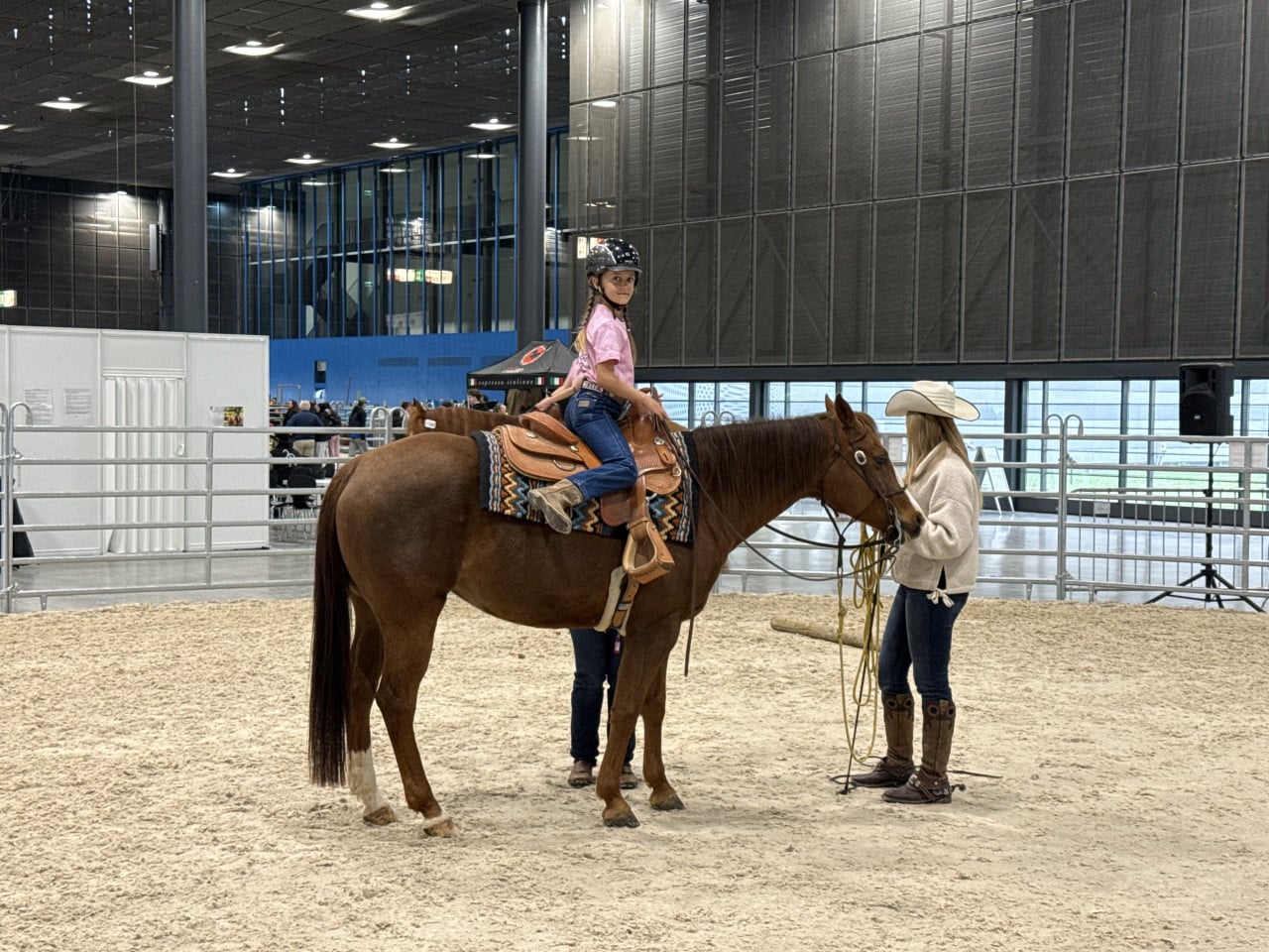 Dívka v jezdecké helmě sedí na hnědém koni v aréně, vedle stojí vodička.