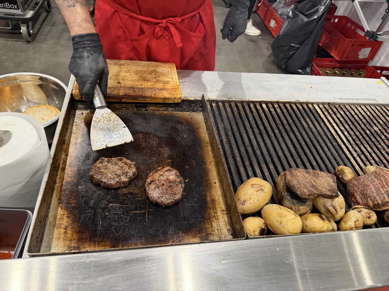 Grilované karbanátky a brambory na stánku, kuchař s obracečkou v červené zástěře