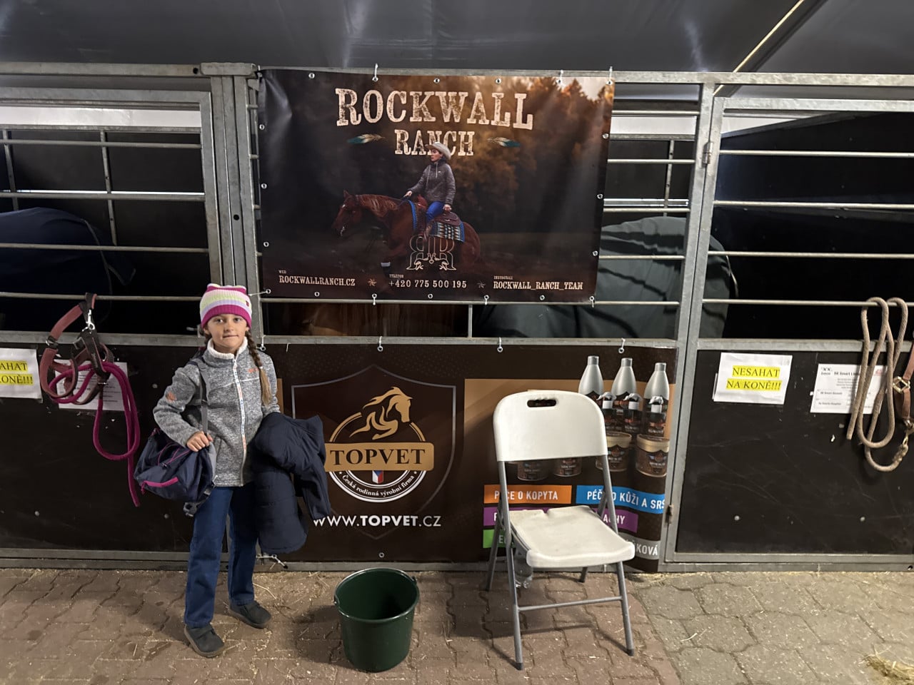 Dívka stojí před stájí s nápisy Rockwall Ranch a Topvet, vedle ohlávky a židle.