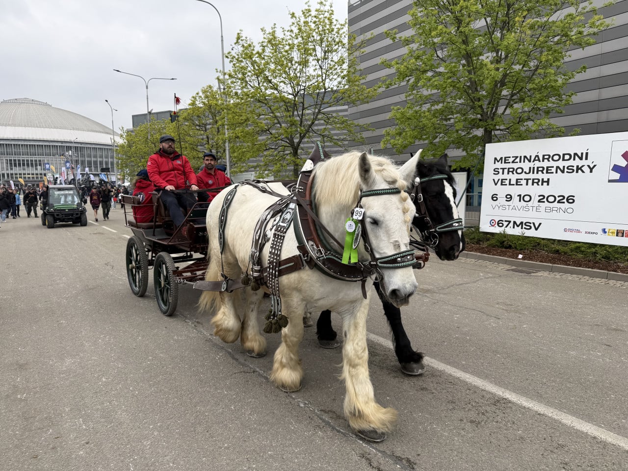 Dvojspřeží koní táhne kočár před brněnským výstavištěm, v pozadí veletržní stánek.