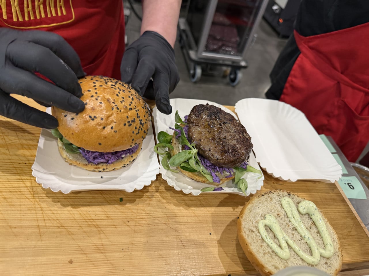 Ruce v černých rukavicích skládají burger s masem, zeleninou a sezamovou bulky.