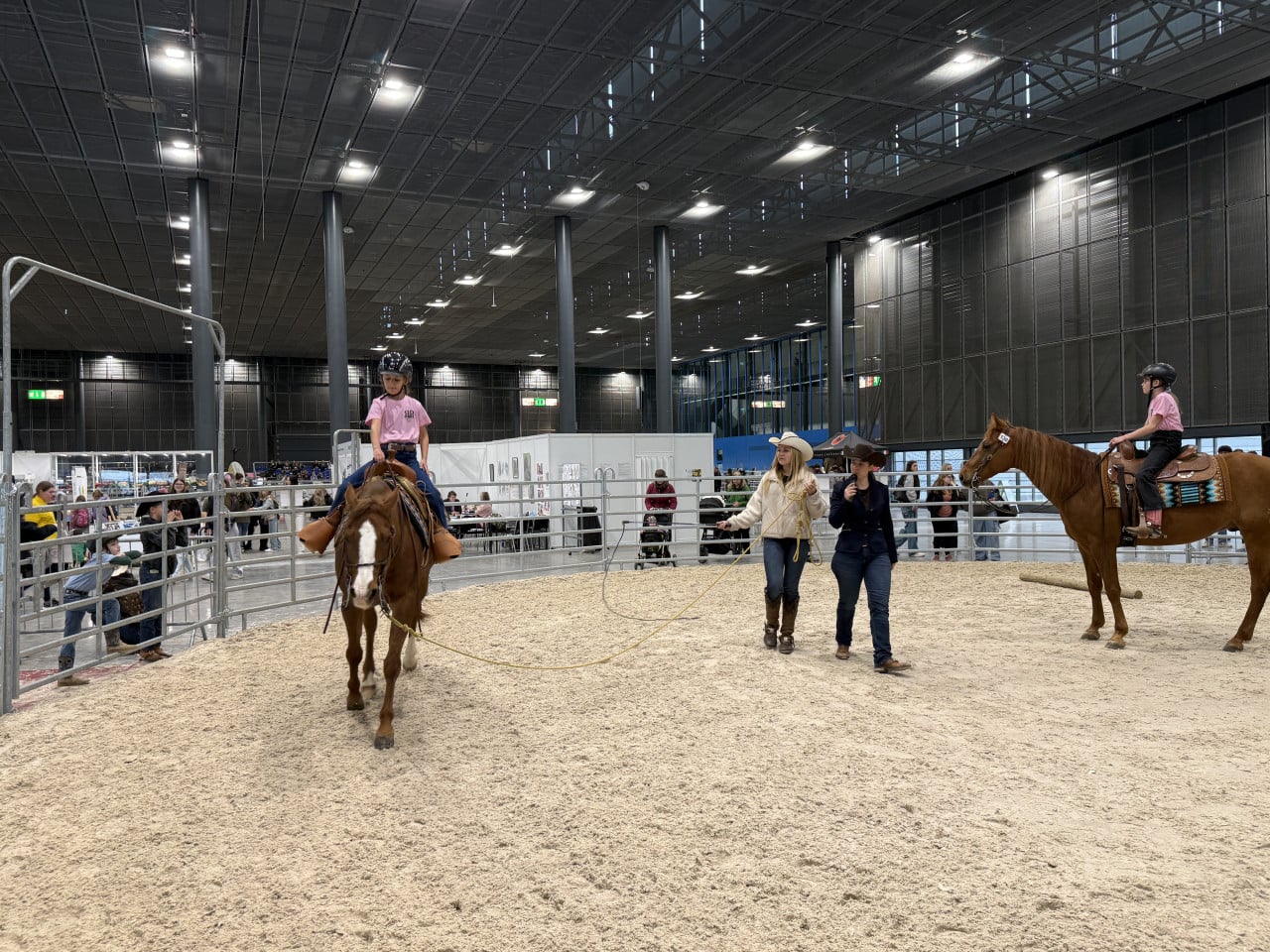 Děti na koních v jezdecké hale, kolem stojí dva vedoucí a diváci.