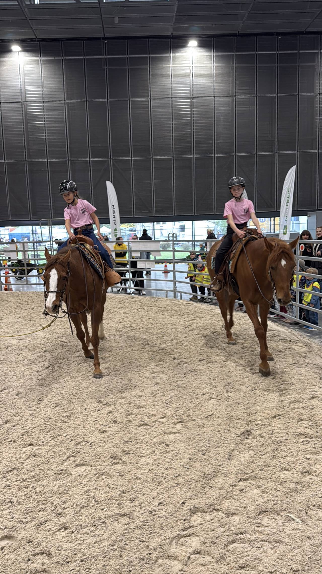 Dvě děti v růžových tričkách jedou na hnědých koních v aréně výstaviště.