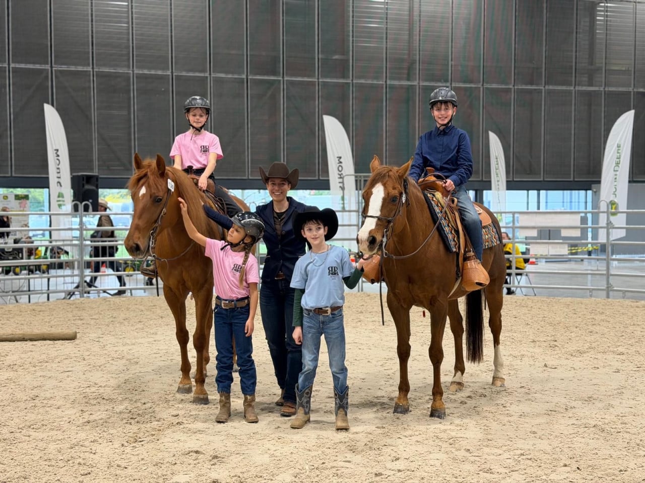 Děti a trenérka pózují s dvěma hnědými koňmi v jezdecké hale