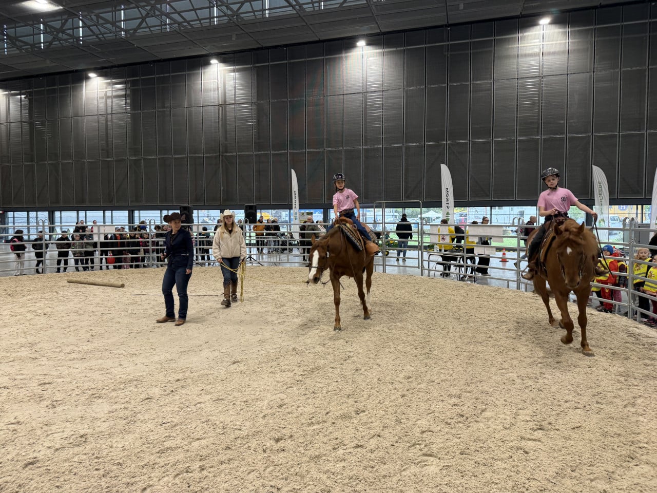 Dvě děti na koních v aréně, vedle nich dva dospělí a sledující publikum.