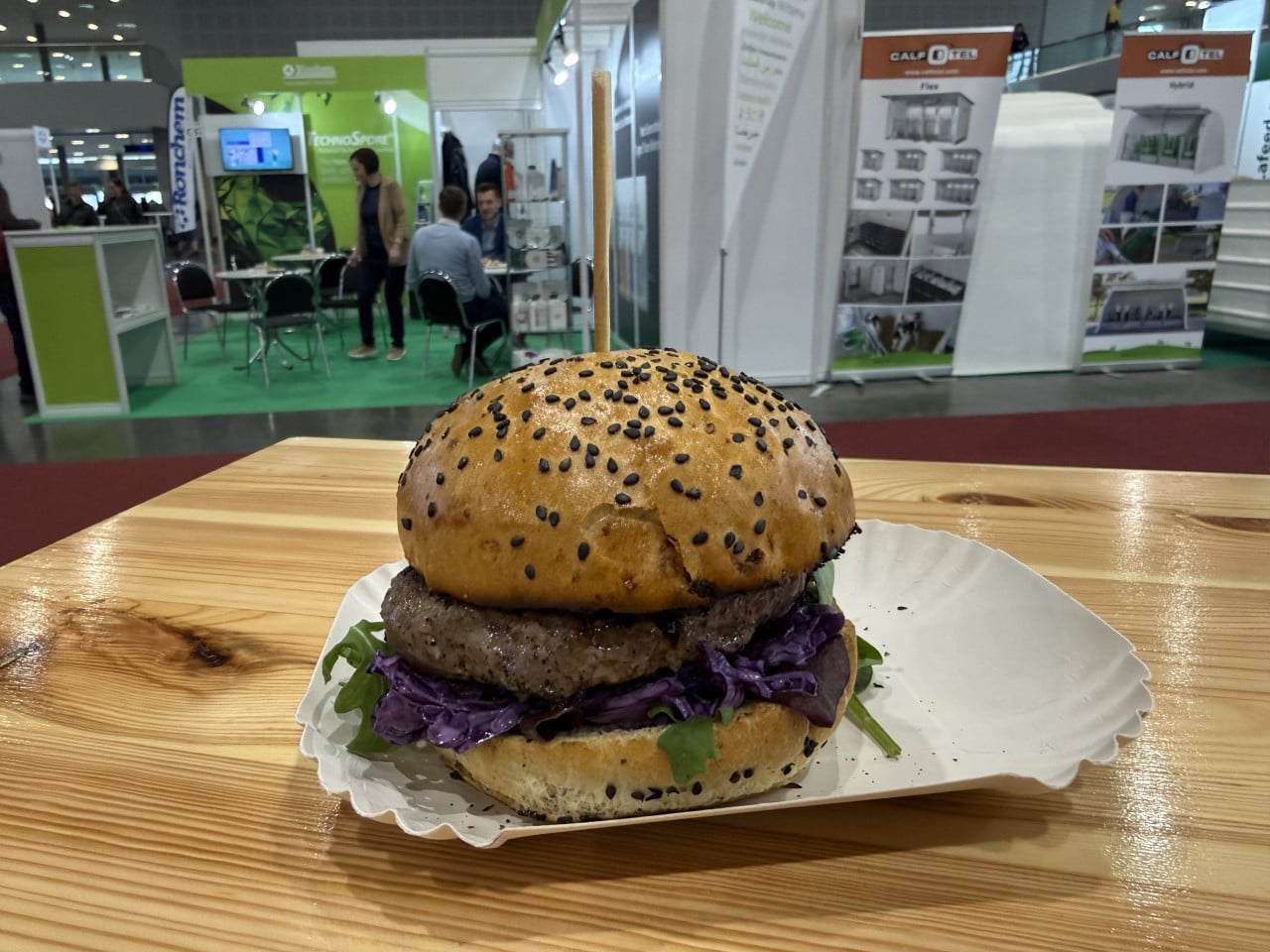 Burger na stole na veletrhu, v pozadí stánky a návštěvníci výstaviště.