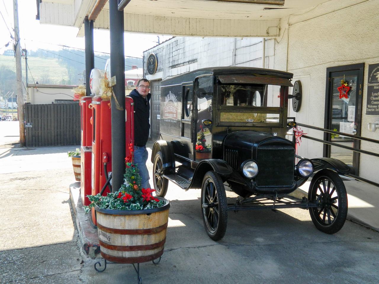 Historický černý pickup u červených stojanů, nápis Lynchburg Hardware, muž u auta.