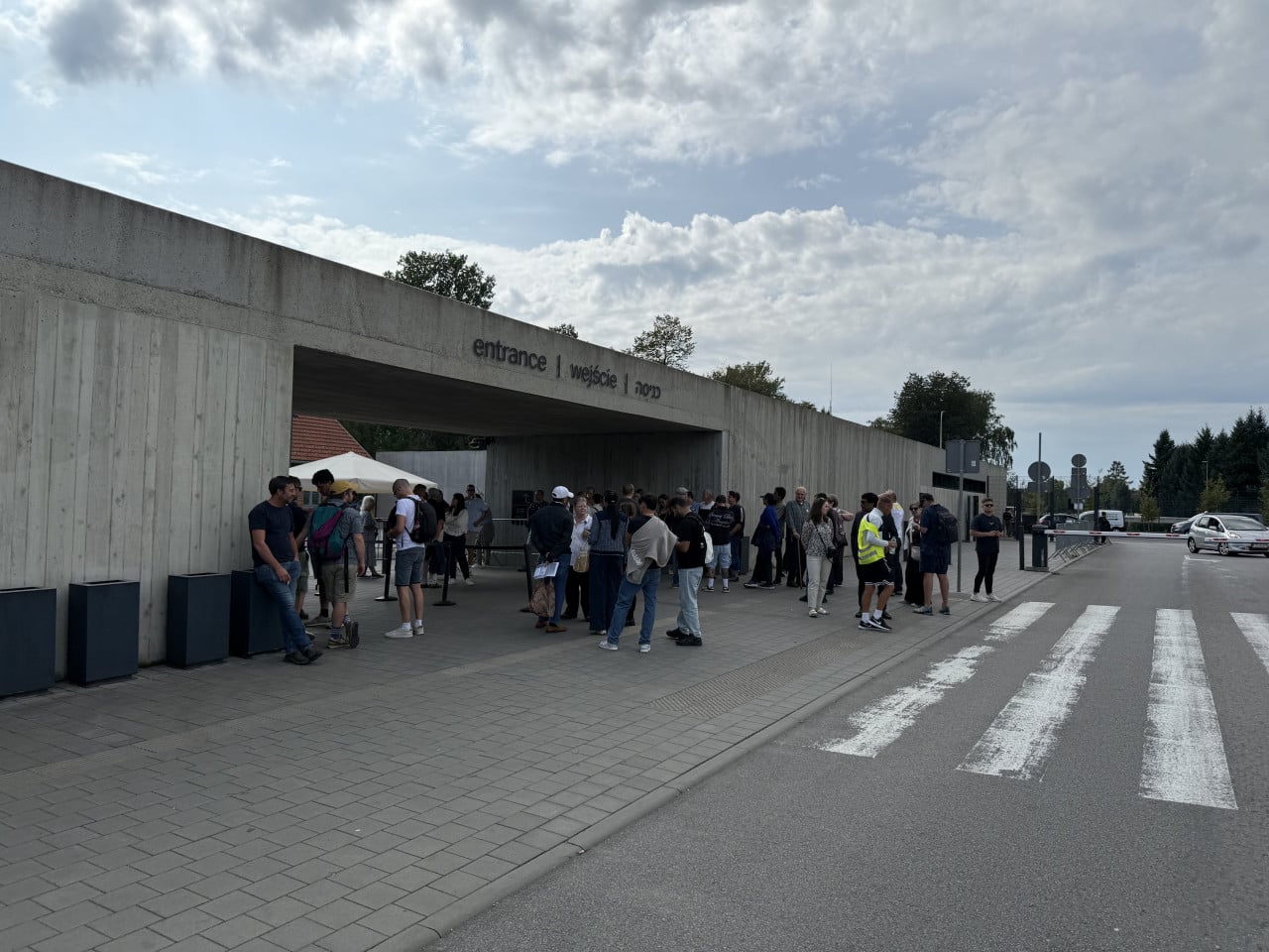 Vstup do Auschwitz-Birkenau s nápisem entrance | wejście | 入り口 a čekajícími návštěvníky