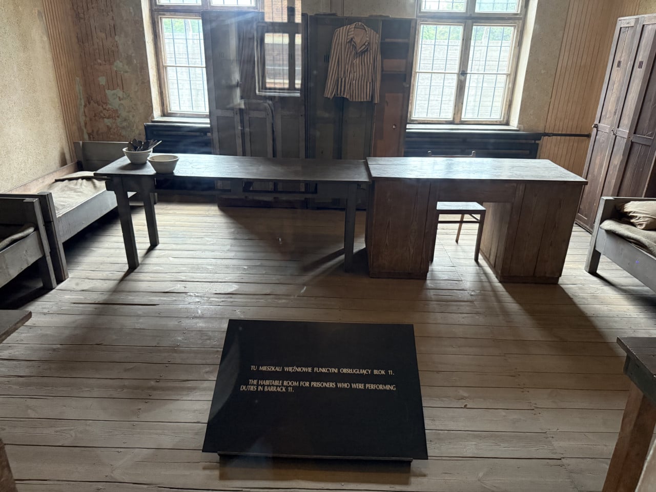 Rekonstrukce místnosti v Auschwitz s postelemi, stolem a pruhovanou vězeňskou košilí.