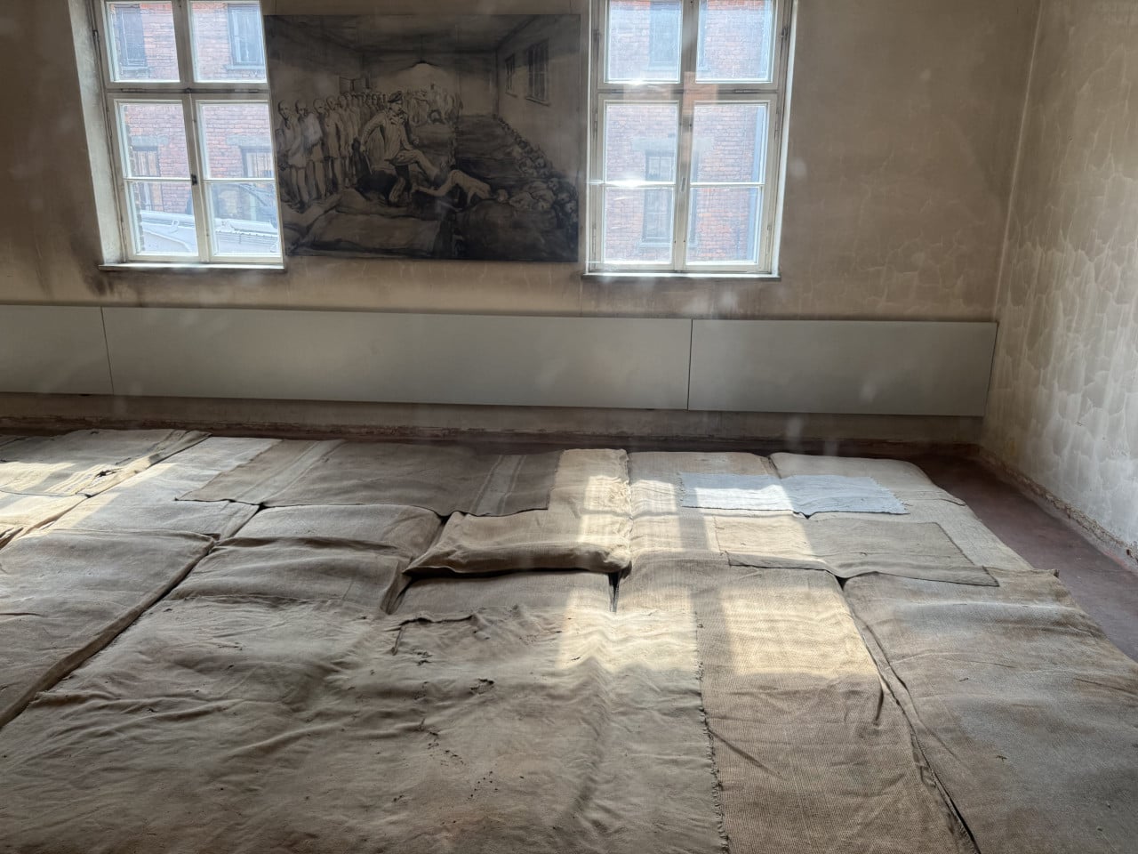 Prázdná místnost s pruhy látky na podlaze a kresbou za oknem v Auschwitz-Birkenau