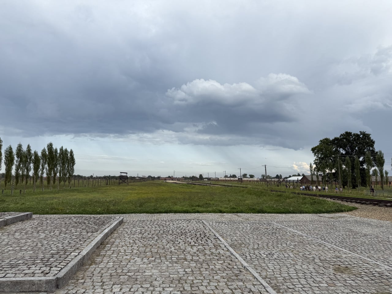 Pohled na rozlehlé pole Birkenau, koleje, věž a dramaticky zataženou oblohu
