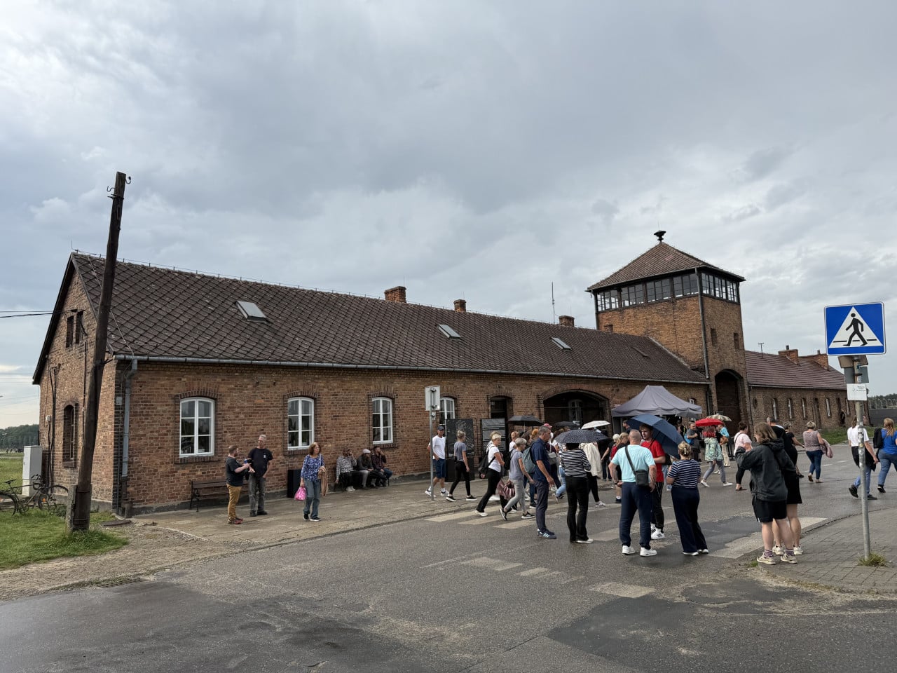 Návštěvníci před cihlovou budovou a strážní věží v Auschwitz-Birkenau pod zataženou oblohou