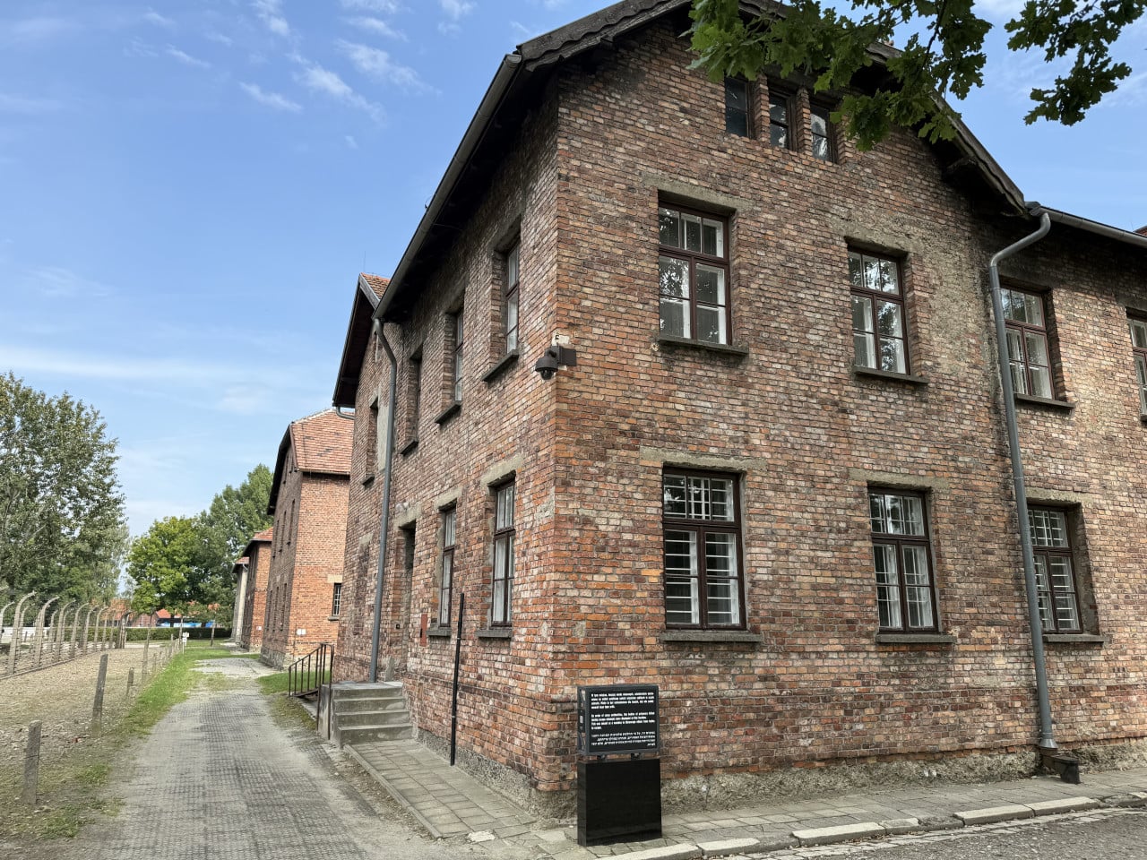Cihlové budovy v Auschwitz-Birkenau pod modrou oblohou, oplocení a informační tabule
