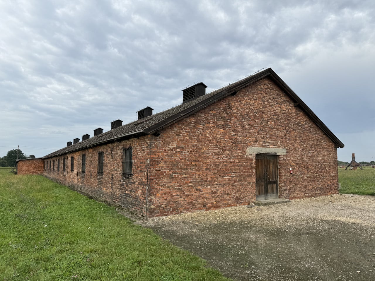 Cihlová budova baráku v Auschwitz-Birkenau pod zataženou oblohou a travnatým polem