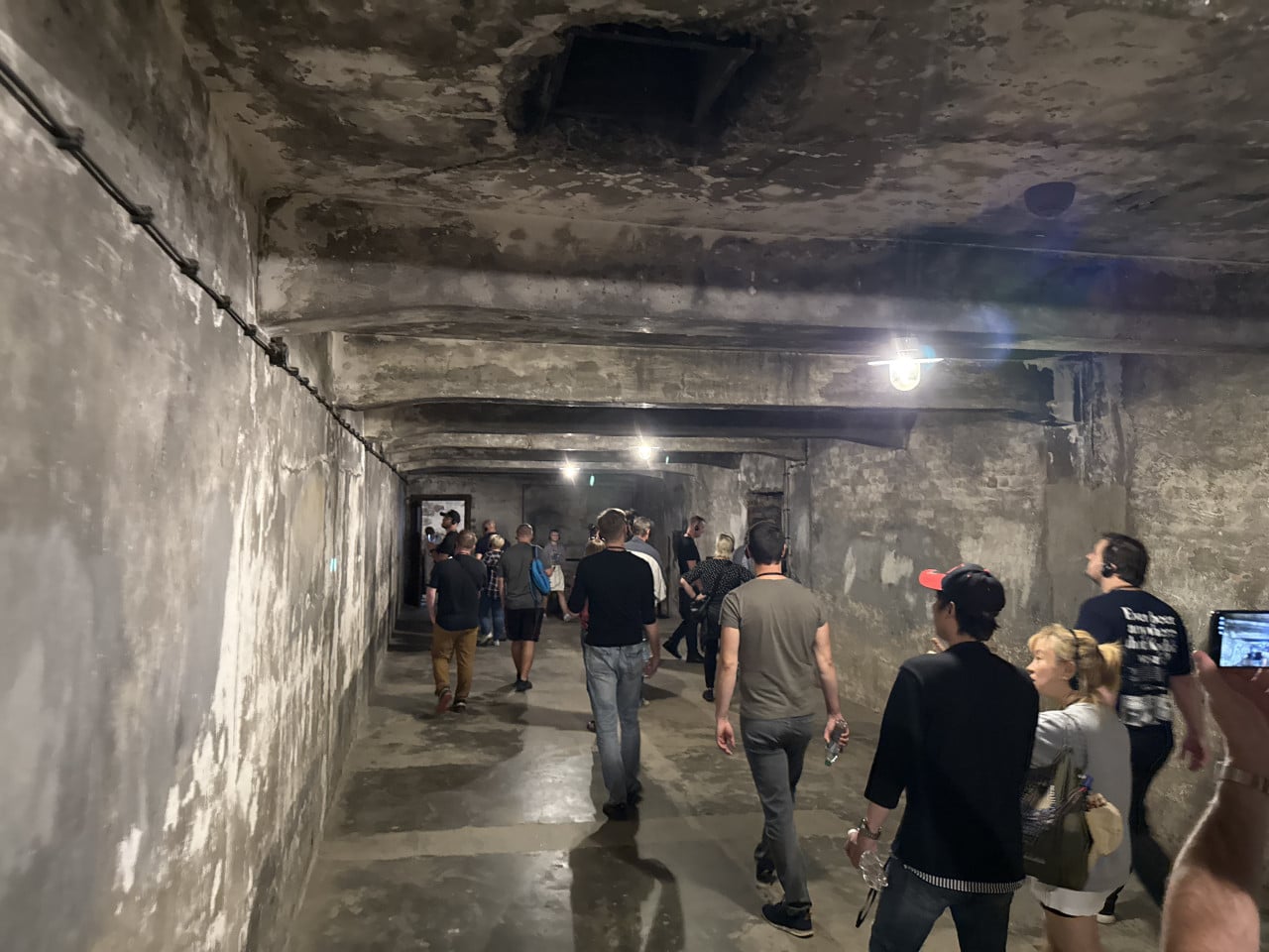 Návštěvníci procházejí šedou betonovou chodbou v Auschwitz s nízkým stropem.