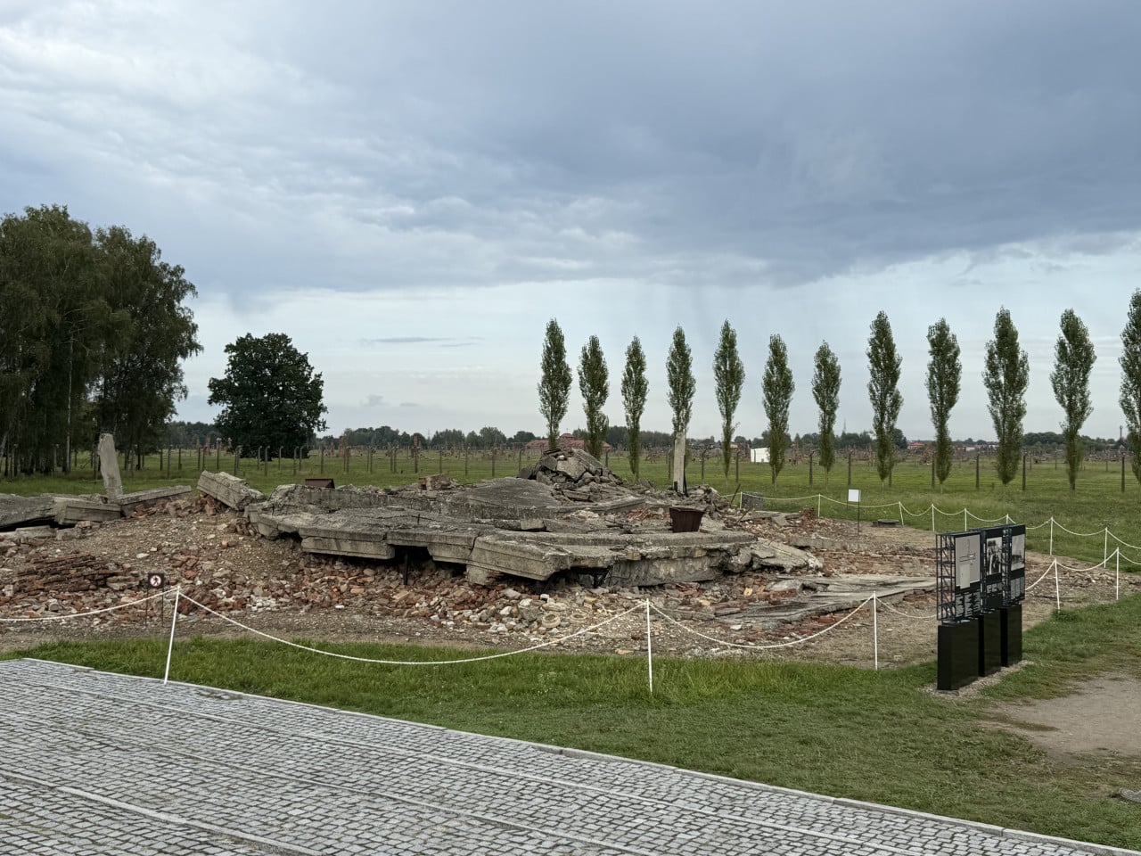 Zřícené betonové základy v Birkenau pod zamračenou oblohou a řadou štíhlých stromů