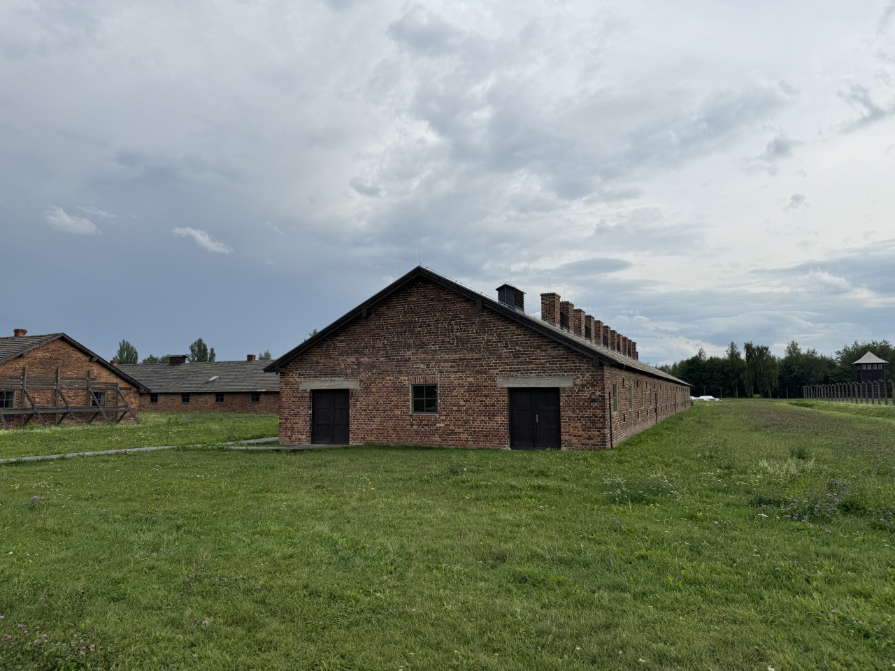 Cihlové baráky v Auschwitz-Birkenau pod zamračenou oblohou, travnaté nádvoří, plot a strážní věž