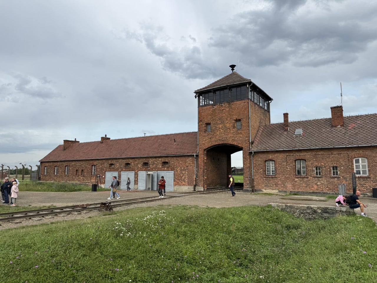 Cihlová brána Auschwitz-Birkenau s kolejemi, návštěvníky a zamračenou oblohou