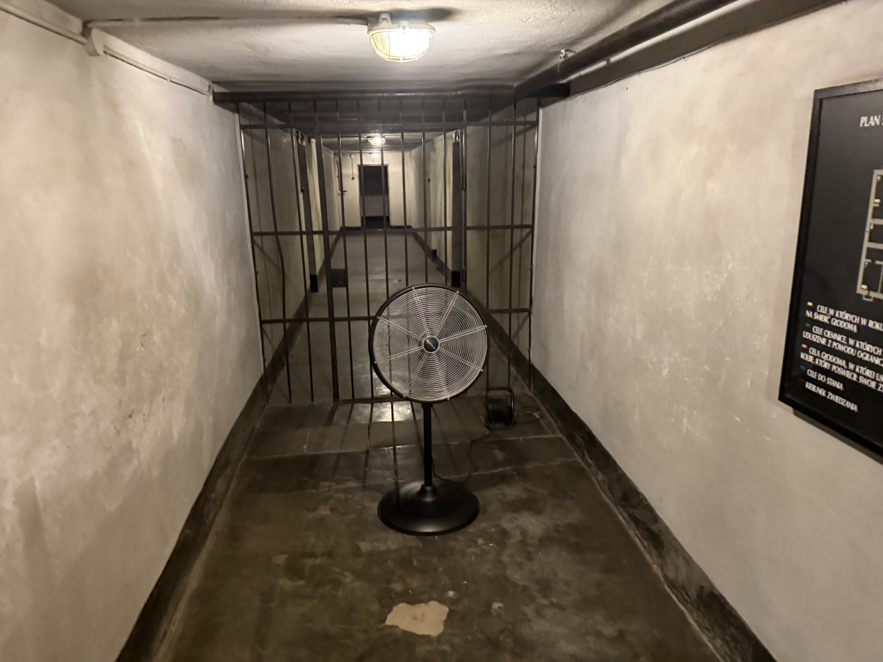Úzká vězeňská chodba s mřížemi, stojacím ventilátorem a tlumeným osvětlením.