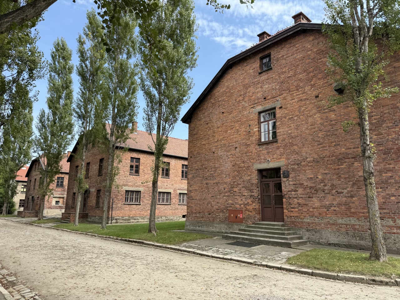 Cihlové budovy Auschwitz-Birkenau lemované stromy pod modrou oblohou