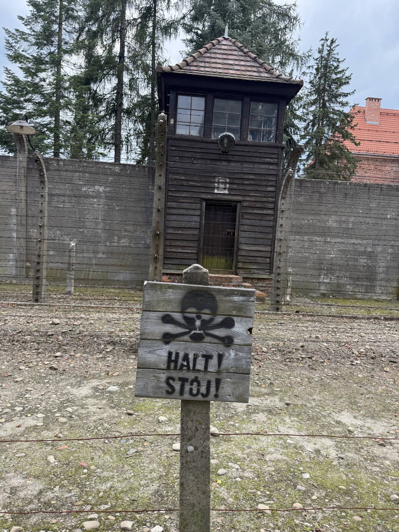 Dřevěná strážní věž za plotem v Osvětimi, před ní cedule s nápisem HAJTY STÓJ!