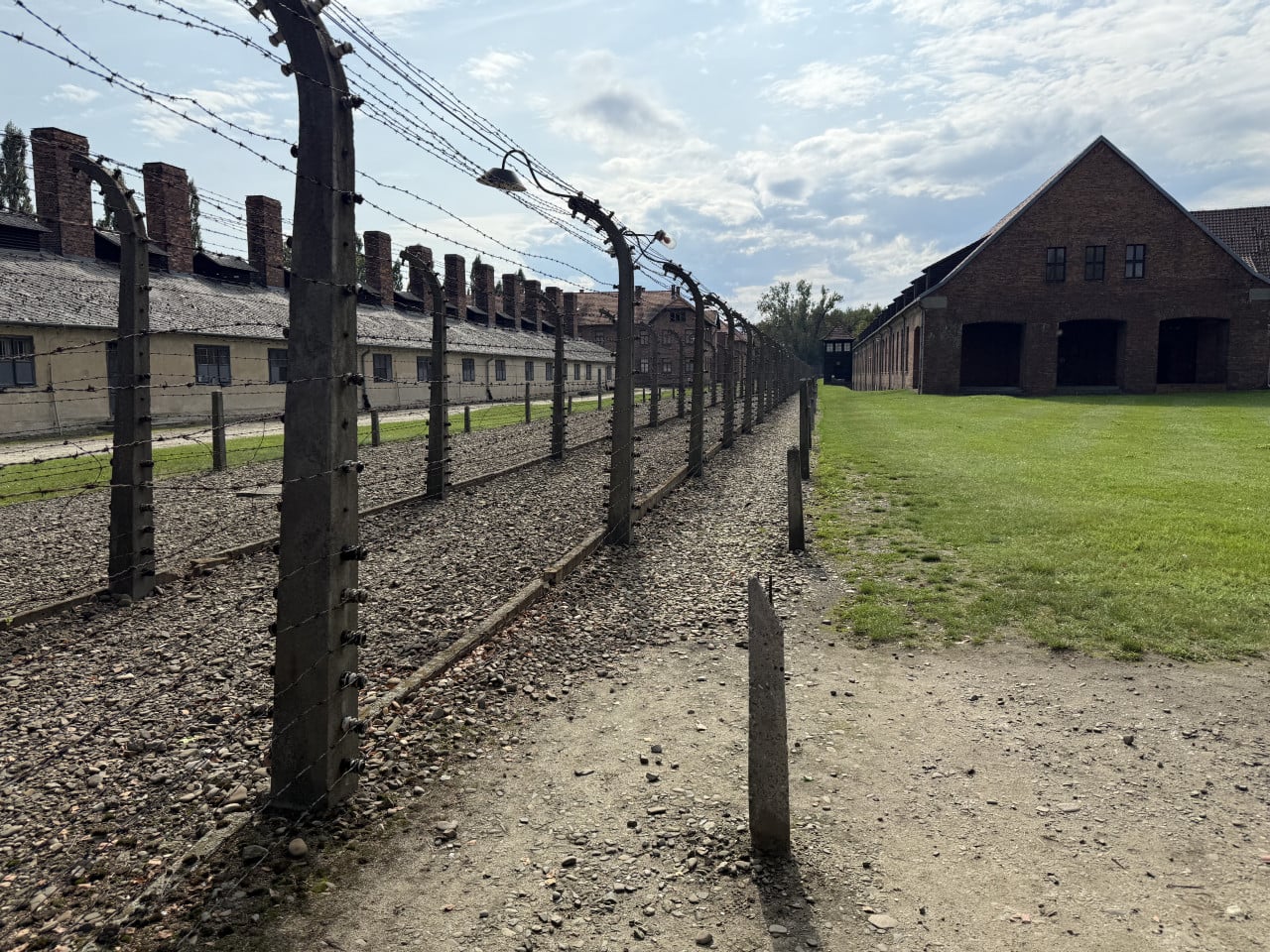 Plot s ostnatým drátem a cihlovými baráky v Auschwitz-Birkenau pod zataženou oblohou