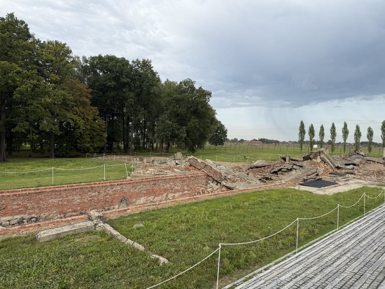 Zříceniny cihlových budov v Birkenau, obehnané provazy, pod zataženou oblohou.