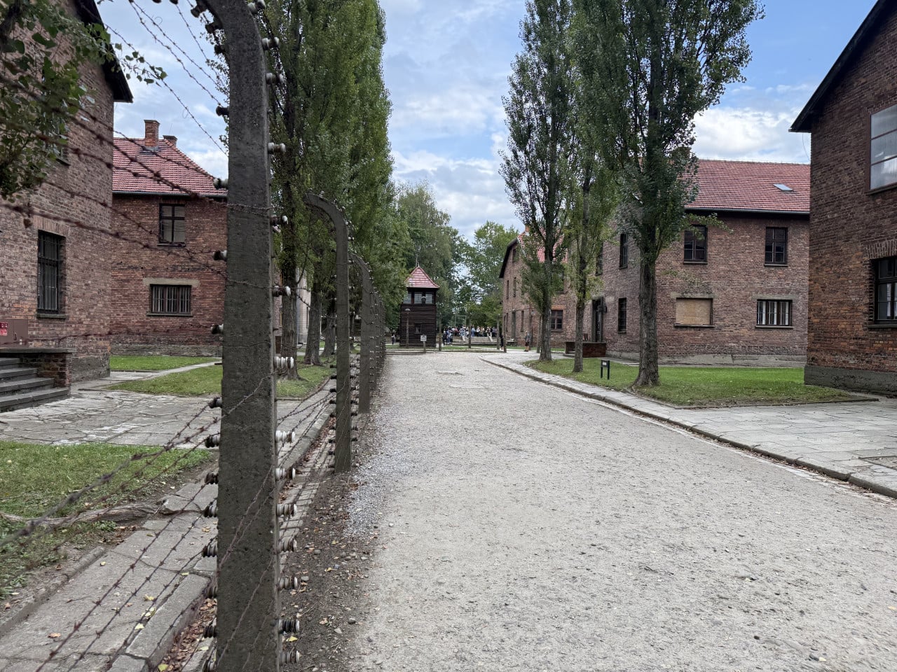 Cesta mezi cihlovými bloky v Auschwitz-Birkenau s ostnatým drátem a strážní věží.