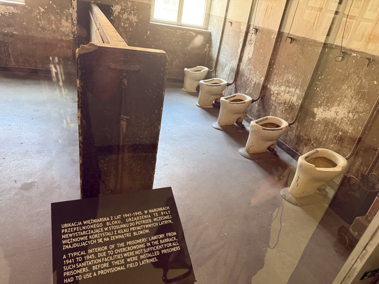 Prostor táborové toalety s řadou záchodů a informační tabulkou Auschwitz-Birkenau