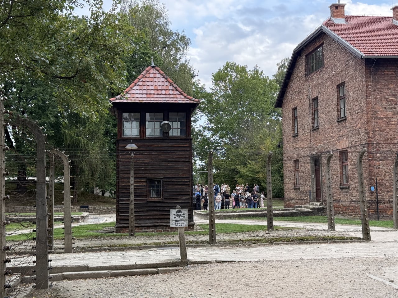 Strážní věž a cihlové budovy v Auschwitz-Birkenau za ostnatým drátem, skupina návštěvníků v pozadí