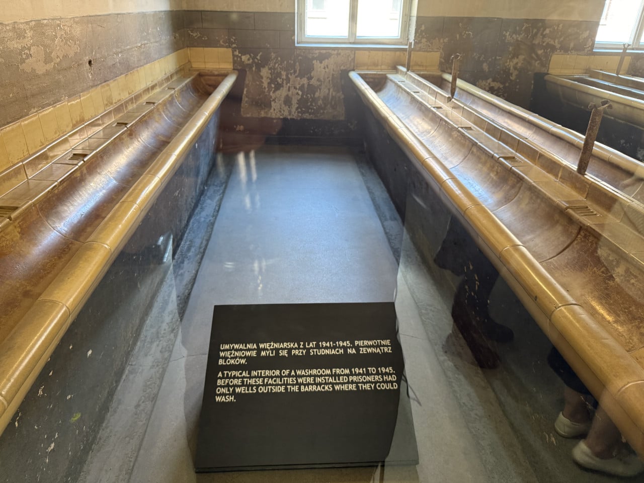 Interiér vězeňské umývárny s betonovými koryty a informačním panelem v Auschwitz-Birkenau